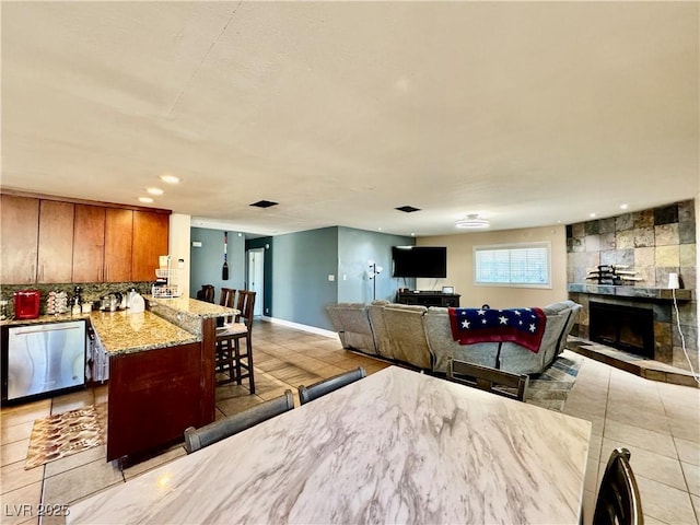 interior space with light tile patterned floors, a kitchen bar, a stone fireplace, dishwasher, and light stone countertops