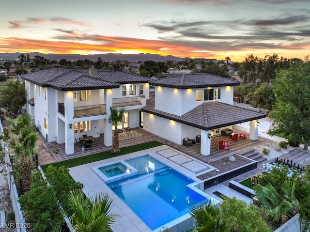 back house at dusk with a pool with hot tub, a patio area, a balcony, an outdoor kitchen, and a bar