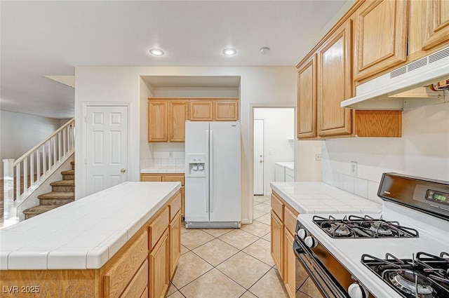 kitchen featuring tile countertops, light tile patterned flooring, light brown cabinetry, gas range, and white fridge with ice dispenser