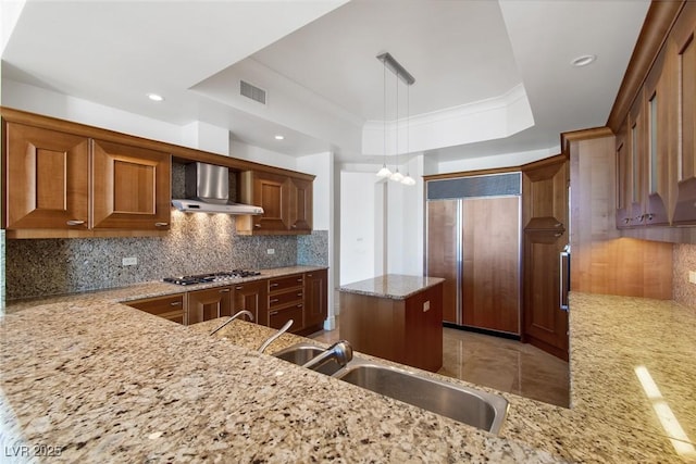 kitchen with decorative light fixtures, a raised ceiling, decorative backsplash, wall chimney exhaust hood, and paneled built in refrigerator