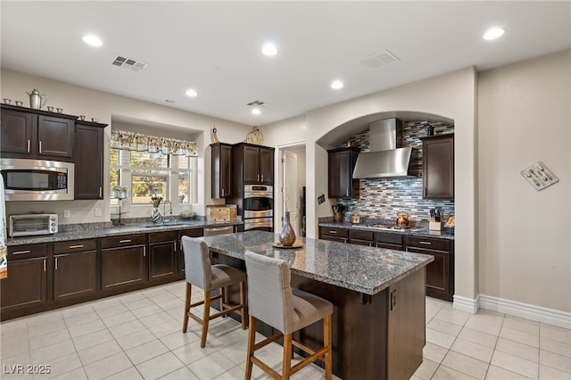 kitchen featuring appliances with stainless steel finishes, sink, dark stone countertops, a center island, and wall chimney range hood