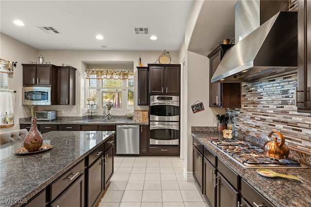 kitchen with wall chimney range hood, light tile patterned floors, sink, appliances with stainless steel finishes, and dark stone countertops