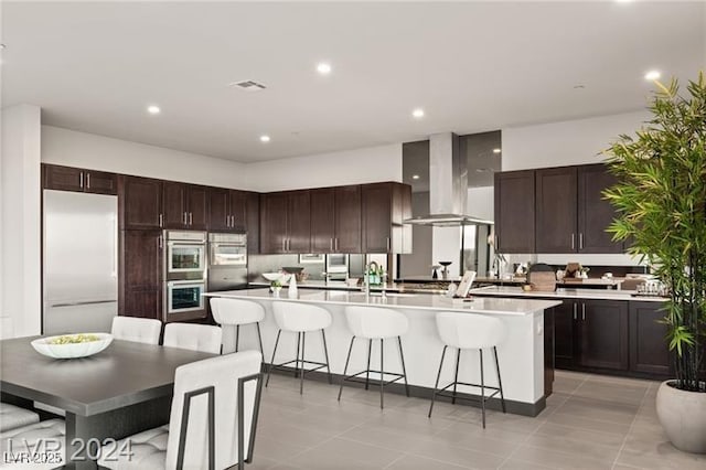 kitchen featuring exhaust hood, light tile patterned flooring, an island with sink, a breakfast bar area, and stainless steel appliances