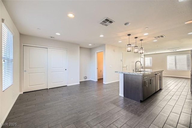 kitchen featuring a center island with sink, sink, dark brown cabinetry, hanging light fixtures, and light stone counters