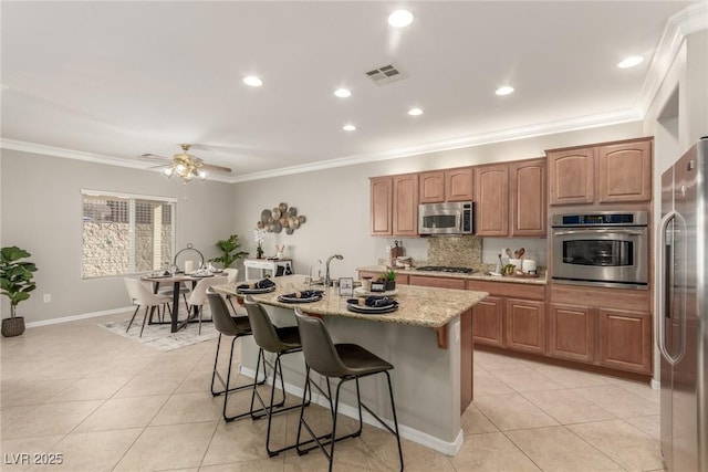 kitchen with appliances with stainless steel finishes, ornamental molding, ceiling fan, a breakfast bar, and a center island with sink