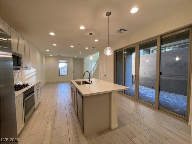 kitchen featuring light hardwood / wood-style floors, a center island with sink, appliances with stainless steel finishes, hanging light fixtures, and sink