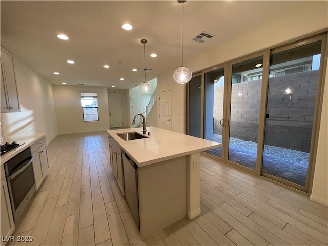 kitchen featuring decorative light fixtures, a center island with sink, sink, light wood-type flooring, and appliances with stainless steel finishes