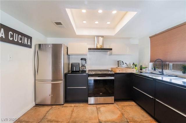 kitchen featuring appliances with stainless steel finishes, white cabinets, wall chimney range hood, sink, and a tray ceiling