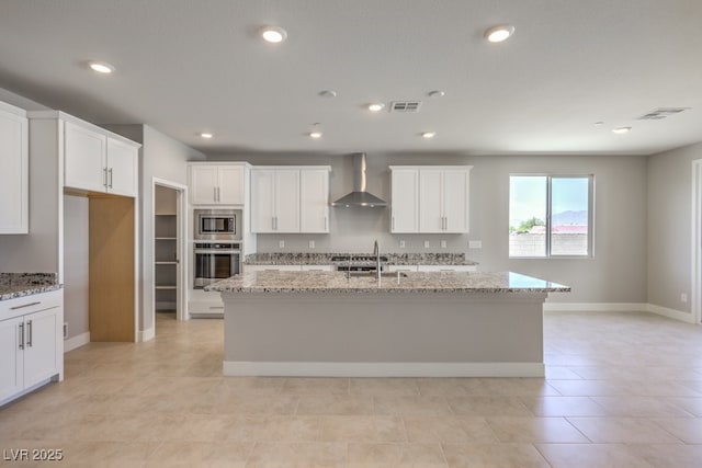 kitchen with appliances with stainless steel finishes, wall chimney exhaust hood, light stone counters, and an island with sink