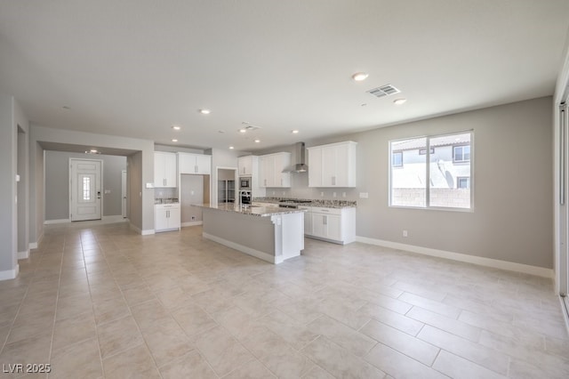 kitchen with light stone countertops, appliances with stainless steel finishes, white cabinetry, wall chimney range hood, and a center island with sink