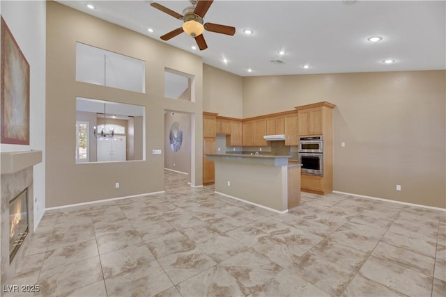 kitchen featuring ceiling fan with notable chandelier, a high ceiling, stainless steel double oven, light brown cabinets, and a premium fireplace