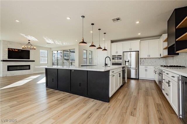 kitchen featuring a premium fireplace, appliances with stainless steel finishes, white cabinetry, an island with sink, and decorative light fixtures