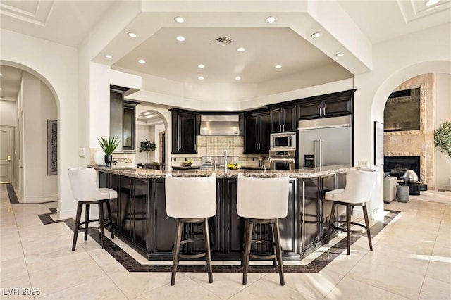 kitchen with a kitchen breakfast bar, built in appliances, light stone counters, a large fireplace, and wall chimney exhaust hood