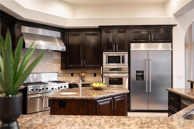 kitchen with sink, dark brown cabinets, built in appliances, light stone counters, and wall chimney exhaust hood