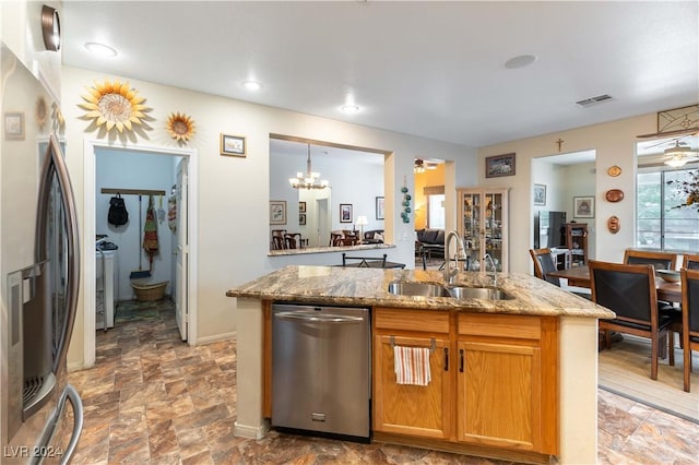 kitchen featuring stainless steel appliances, an inviting chandelier, sink, light stone counters, and a center island with sink