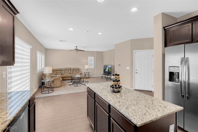 kitchen featuring ceiling fan, a center island, stainless steel appliances, dark brown cabinets, and light stone counters