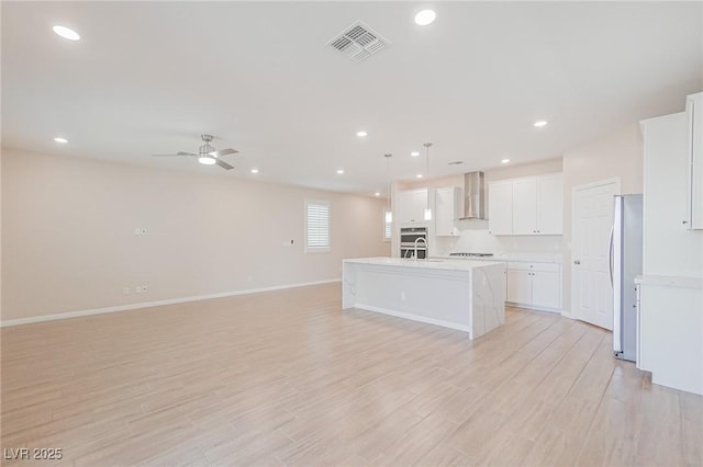 kitchen featuring stainless steel refrigerator, white cabinetry, hanging light fixtures, an island with sink, and wall chimney exhaust hood