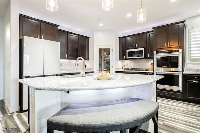 kitchen featuring sink, a kitchen island with sink, hanging light fixtures, stainless steel appliances, and dark brown cabinetry