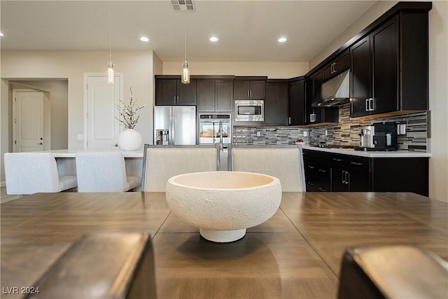 kitchen featuring exhaust hood, stainless steel appliances, decorative backsplash, dark brown cabinets, and hanging light fixtures