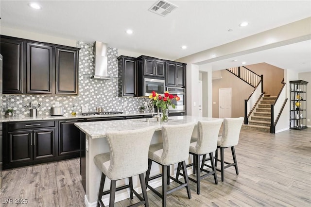 kitchen featuring stainless steel appliances, wall chimney exhaust hood, light hardwood / wood-style flooring, and a center island with sink