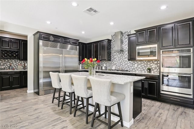 kitchen with light hardwood / wood-style flooring, wall chimney range hood, built in appliances, a center island with sink, and a breakfast bar area