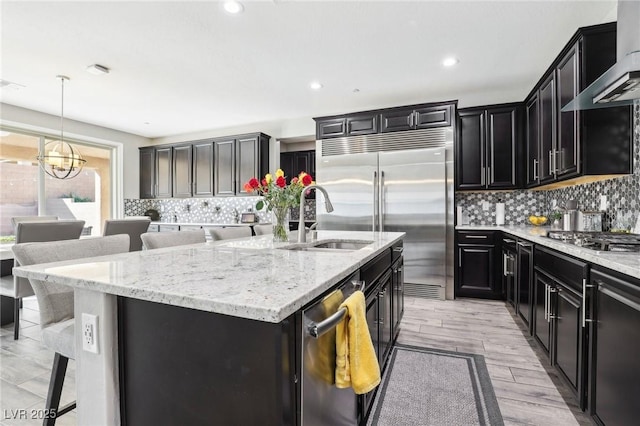 kitchen featuring a center island with sink, stainless steel appliances, a kitchen breakfast bar, wall chimney exhaust hood, and sink