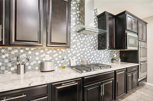 kitchen featuring light stone countertops, dark brown cabinetry, wall chimney exhaust hood, stainless steel appliances, and tasteful backsplash