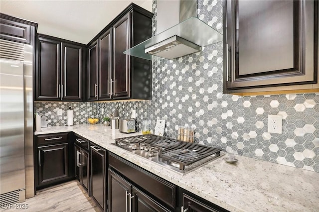 kitchen with backsplash, dark brown cabinetry, appliances with stainless steel finishes, wall chimney exhaust hood, and light stone counters