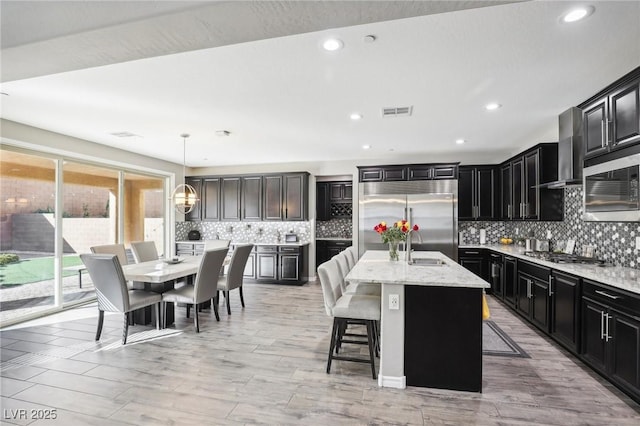 kitchen with a center island with sink, stainless steel appliances, wall chimney exhaust hood, a kitchen breakfast bar, and pendant lighting