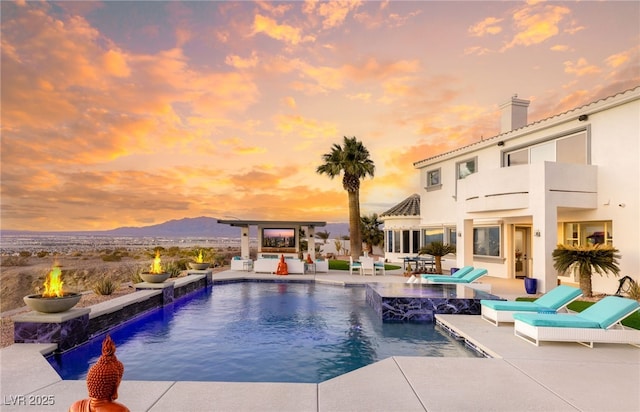 pool at dusk with a mountain view, a patio area, pool water feature, and an outdoor fire pit