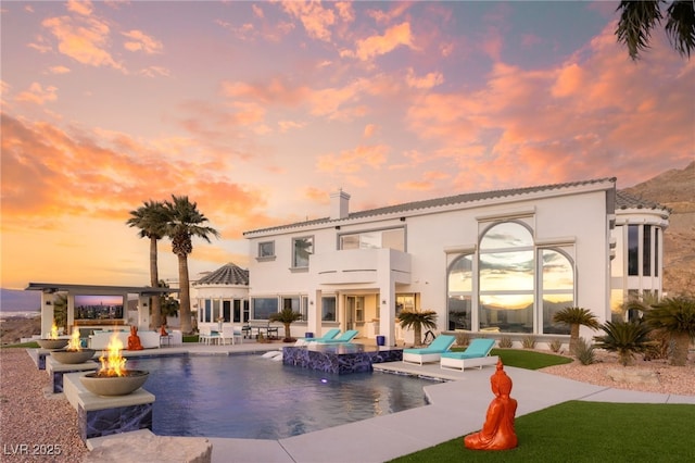 back house at dusk featuring a balcony, a patio area, and a fire pit