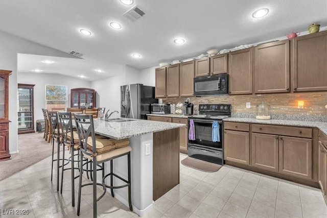 kitchen with vaulted ceiling, a center island with sink, black appliances, a kitchen bar, and light stone counters