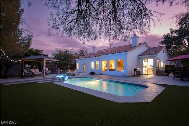 pool at dusk featuring a patio area, a gazebo, grilling area, a lawn, and an in ground hot tub