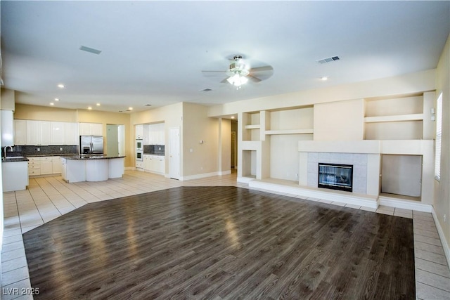 unfurnished living room with a fireplace, sink, built in shelves, light hardwood / wood-style flooring, and ceiling fan