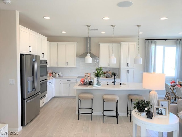 kitchen featuring pendant lighting, white cabinetry, a kitchen island with sink, stainless steel appliances, and wall chimney exhaust hood