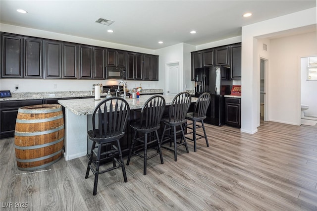 kitchen with light hardwood / wood-style floors, black appliances, a center island with sink, light stone countertops, and a kitchen breakfast bar