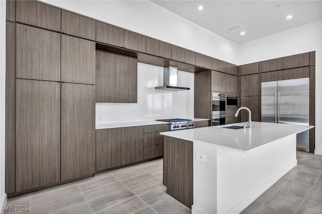 kitchen with sink, a kitchen island with sink, light tile patterned floors, stainless steel appliances, and wall chimney exhaust hood