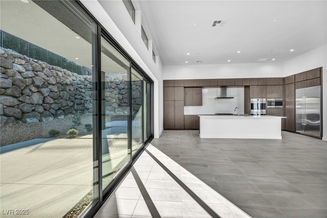 kitchen featuring ventilation hood, stainless steel built in refrigerator, an island with sink, sink, and dark brown cabinetry