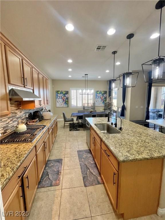 kitchen with light stone countertops, hanging light fixtures, a kitchen island with sink, sink, and stainless steel appliances