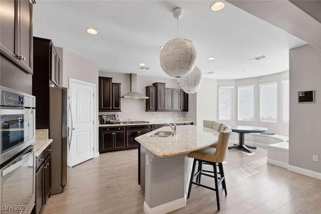 kitchen featuring decorative light fixtures, wall chimney range hood, an island with sink, sink, and light stone counters