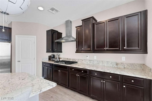 kitchen with black gas stovetop, wall chimney range hood, light hardwood / wood-style floors, stainless steel refrigerator, and dark brown cabinetry