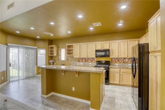 kitchen featuring a center island with sink, light brown cabinetry, light stone counters, a breakfast bar, and black appliances