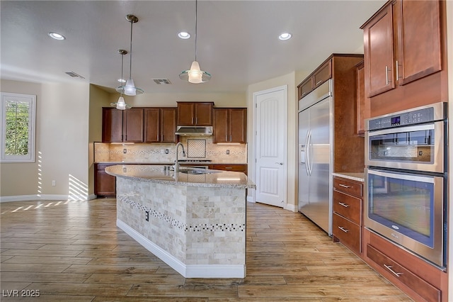 kitchen with light hardwood / wood-style floors, a center island with sink, appliances with stainless steel finishes, decorative backsplash, and decorative light fixtures