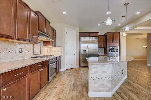 kitchen featuring an island with sink, appliances with stainless steel finishes, light wood-type flooring, hanging light fixtures, and light stone countertops