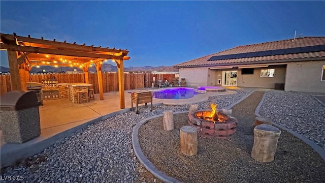 pool at dusk with a patio area, a pergola, a fire pit, exterior kitchen, and an outdoor bar