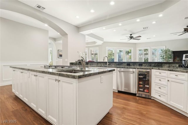 kitchen featuring stainless steel dishwasher, an island with sink, white cabinets, dark stone counters, and beverage cooler