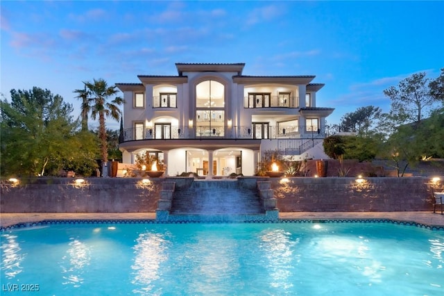 back house at dusk featuring a balcony, a patio area, a fenced in pool, and pool water feature