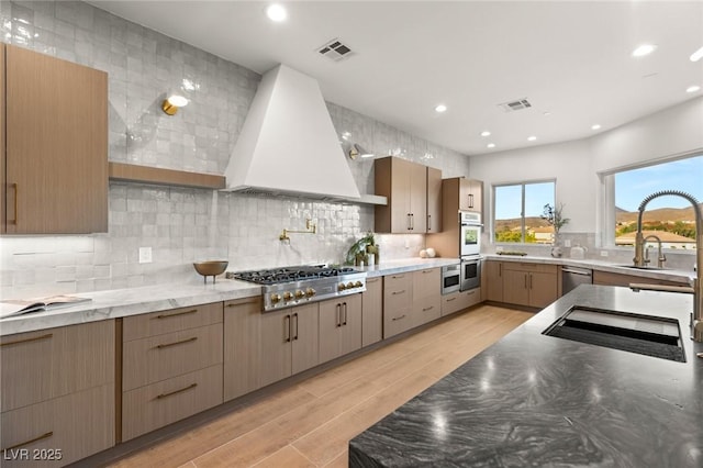 kitchen featuring custom exhaust hood, appliances with stainless steel finishes, decorative backsplash, light hardwood / wood-style flooring, and stone countertops