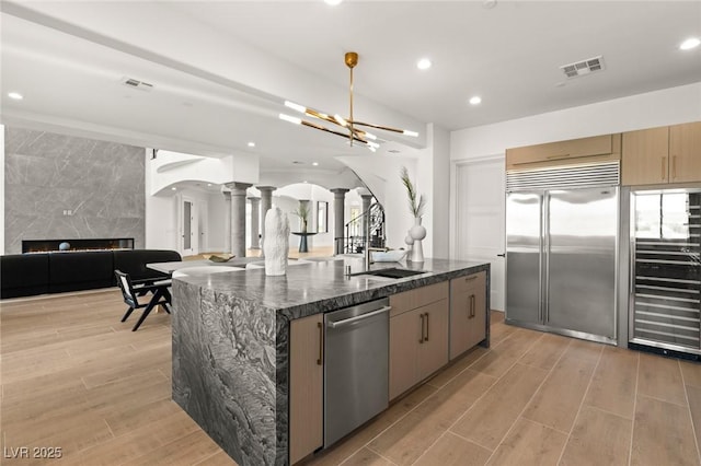 kitchen with sink, an island with sink, ornate columns, and appliances with stainless steel finishes