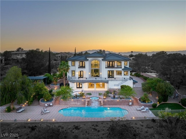 back house at dusk with a balcony, a patio area, pool water feature, and a swimming pool with hot tub